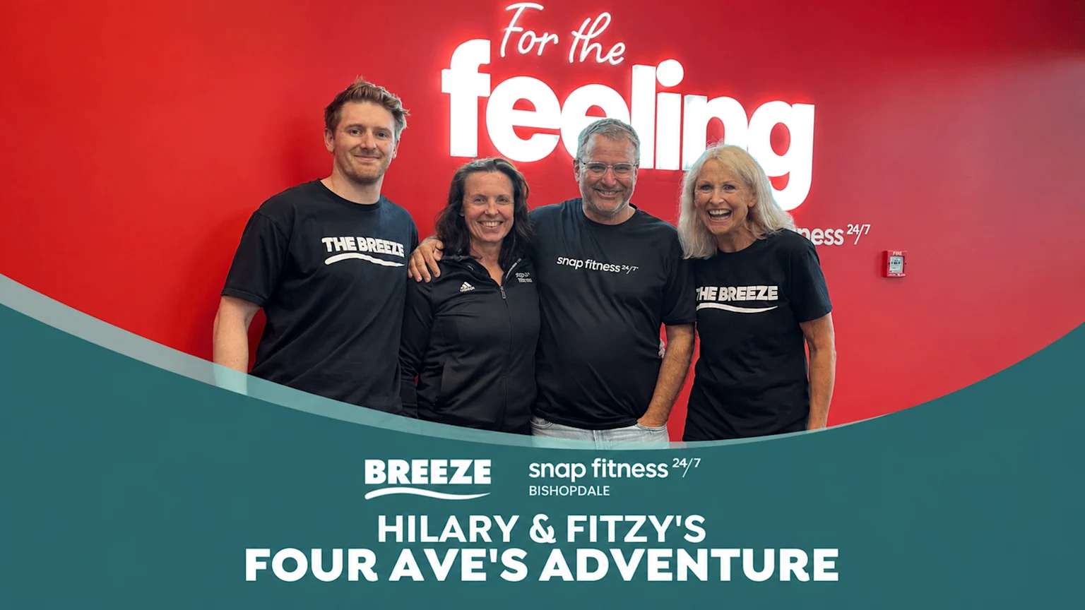 Four smiling people, two men and two women, stand in front of a red wall displaying "For the feeling" and "snap fitness 24/7" text, wearing branded shirts for "THE BREEZE" and "snap fitness".