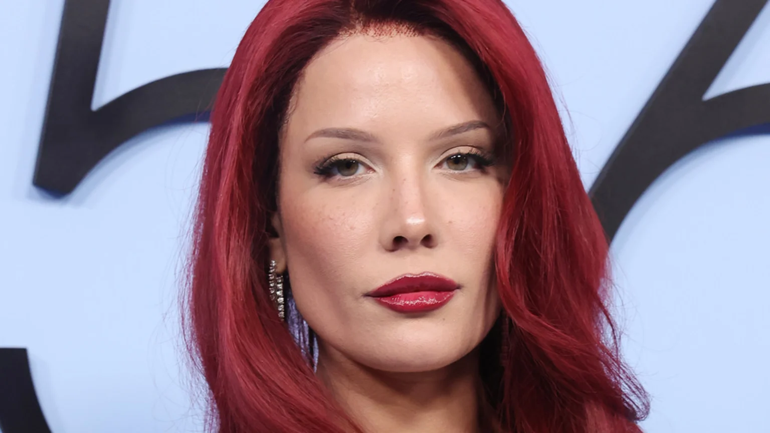 Close-up of a person with long, bright red hair, fair skin, glossy red lipstick, winged eyeliner, and a sparkling earring, against a light blue background.