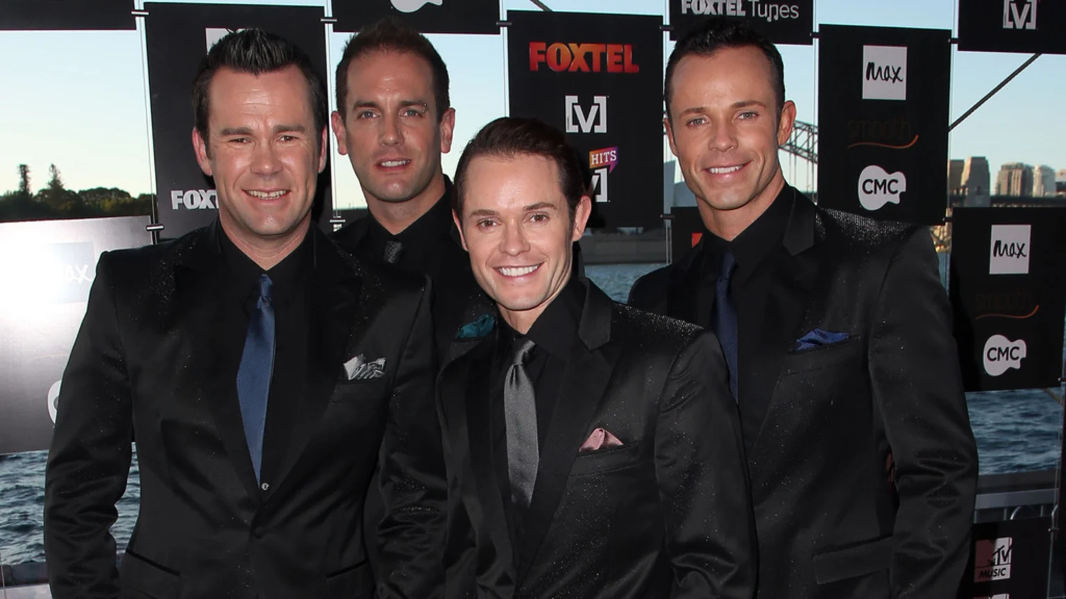 Four men in shimmering black suits and black shirts with colorful ties smile at the camera, standing before a branded backdrop and a city skyline.