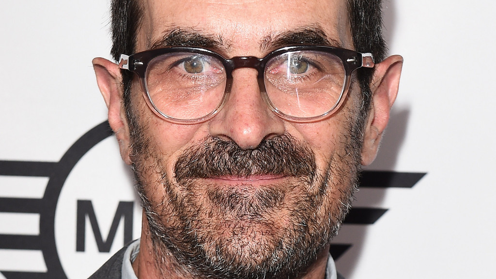 Close-up of actor Ty Burrell wearing dark-rimmed glasses, with a salt-and-pepper mustache and beard, smiling gently against a white background with a partial black "MINI" logo.