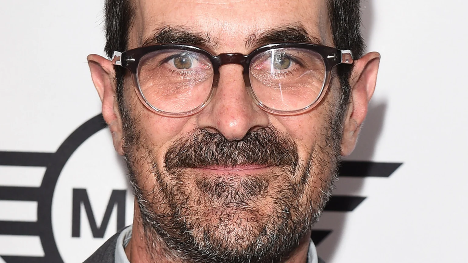 Close-up of actor Ty Burrell wearing dark-rimmed glasses, with a salt-and-pepper mustache and beard, smiling gently against a white background with a partial black "MINI" logo.