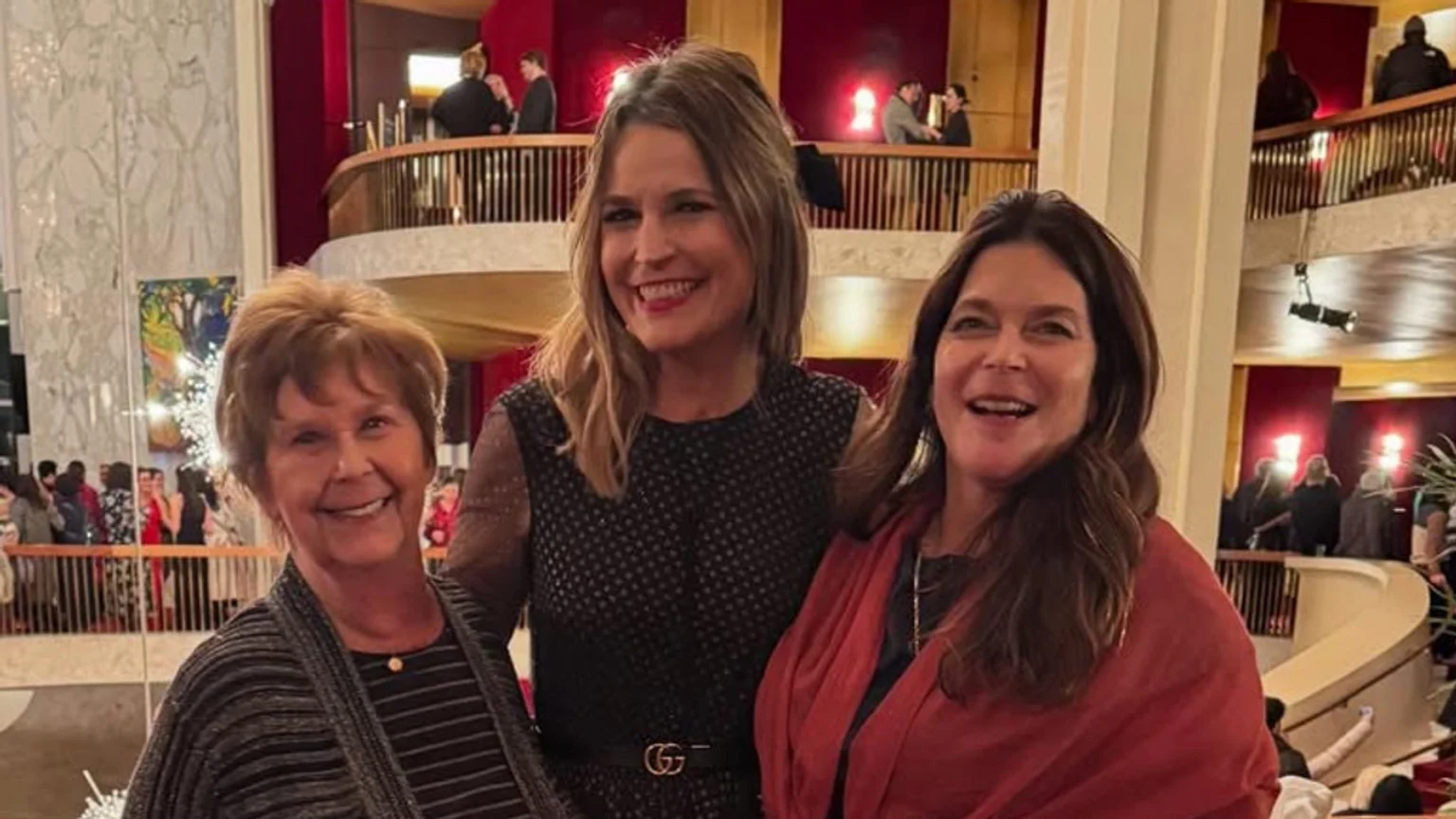 Three women pose and smile for a photo in a dimly lit, grand theater lobby with multiple levels, dark wooden railings, red wall panels, and a white marble-patterned wall. The center woman wears a black belt with a gold 'G' buckle.