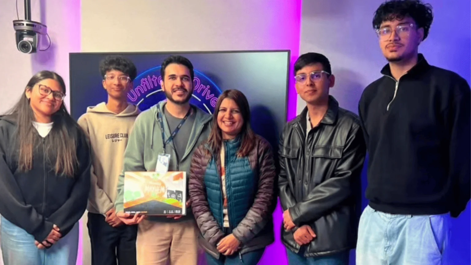 Six smiling people, including a man holding a 'Money Mayhem' box, stand in front of a screen displaying 'Unfiltered Drive.'