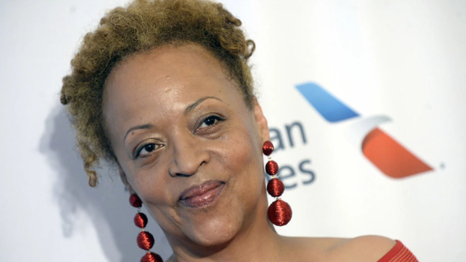 A woman with light brown curly hair and red dangling earrings smiles faintly at the camera, with a blurred American Airlines logo visible in the white background.