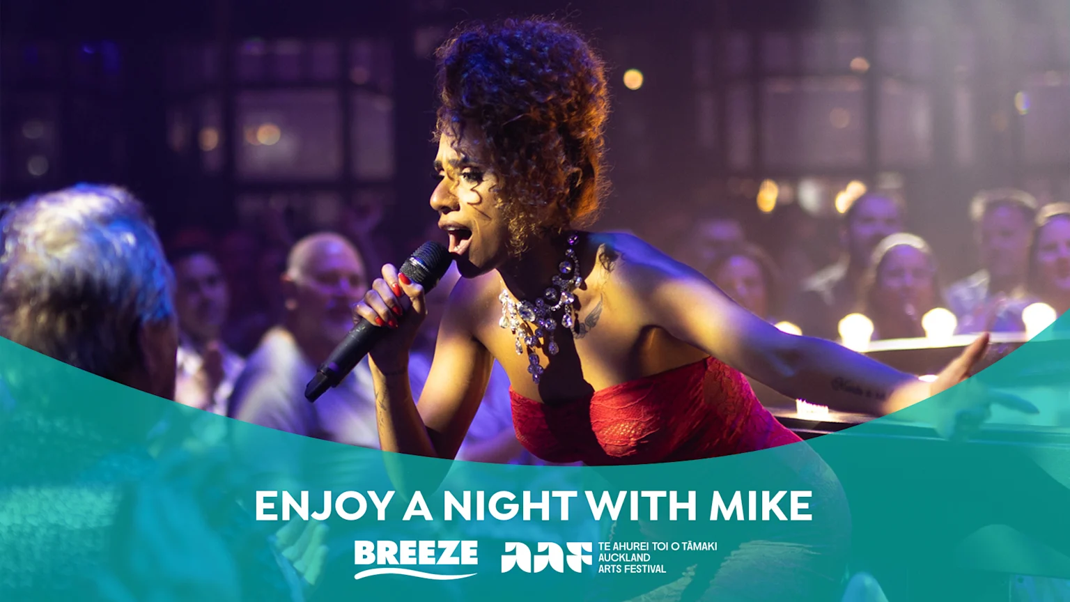 A female singer in a red strapless dress and multi-tiered silver necklace performs on a dimly lit concert hall stage, holding a black microphone. A tattoo on her right forearm is visible. The blurred audience is illuminated by ambient lighting, with event branding from Auckland Arts Festival overlaying the scene.