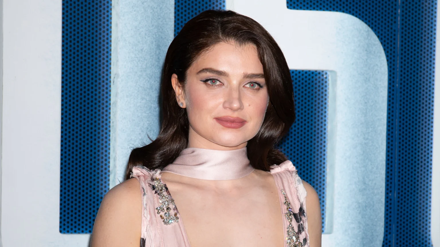 A woman with dark, wavy hair and light eyes looks forward, wearing a pink dress with jeweled straps and a choker, against a blue perforated background.