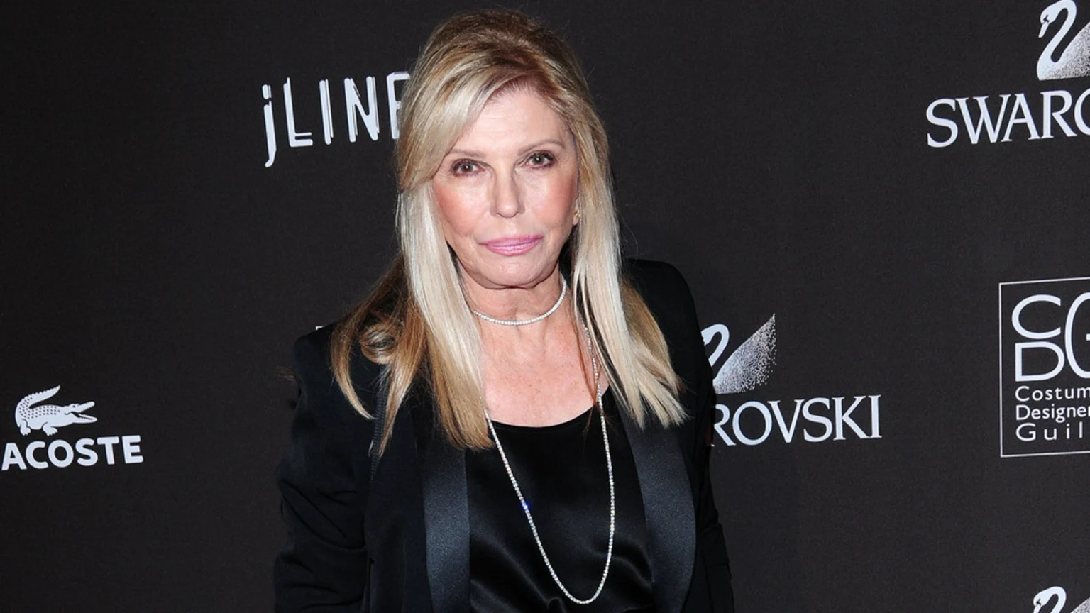 A blonde woman with pink lipstick and diamond necklaces wears a black top and jacket, standing in front of a black backdrop with logos.