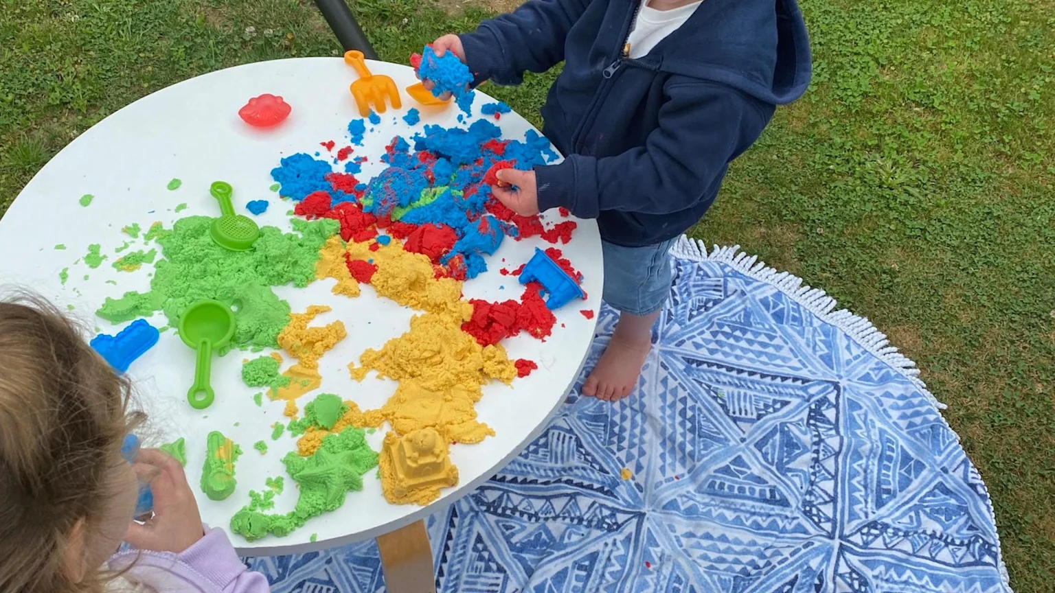 Two children play with colorful kinetic sand and small toys on a white table outdoors, with a blue and white patterned blanket on the grass.