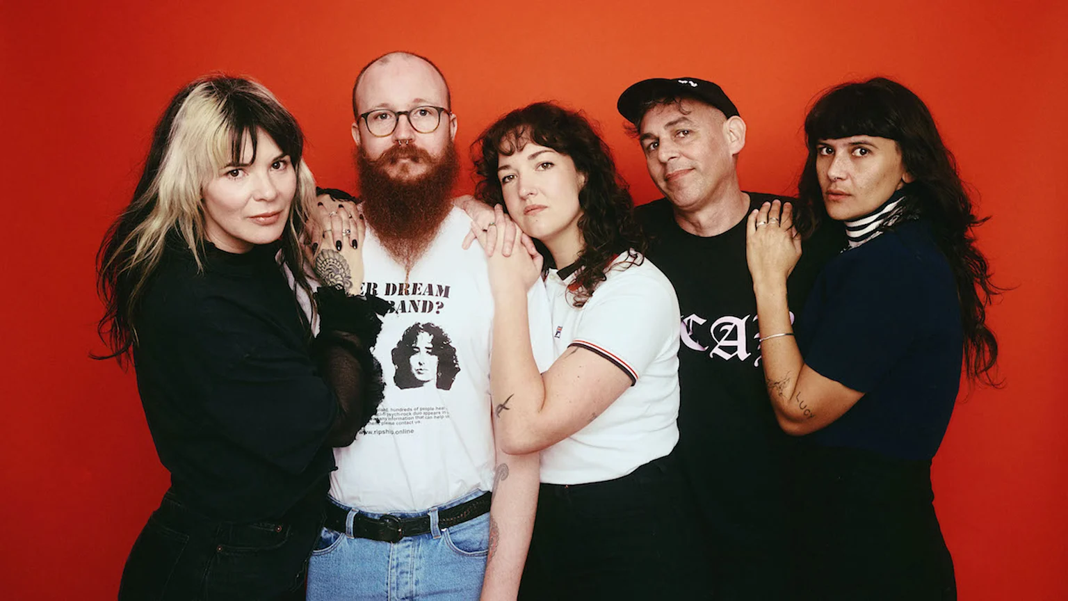A group of five adults, three women and two men, stand against a red background; the central man has a red beard and glasses, and some individuals display tattoos.