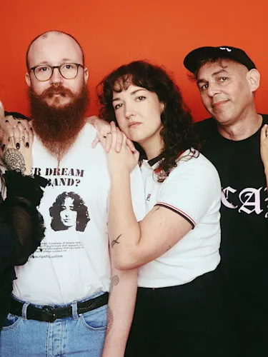 A group of five adults, three women and two men, stand against a red background; the central man has a red beard and glasses, and some individuals display tattoos.