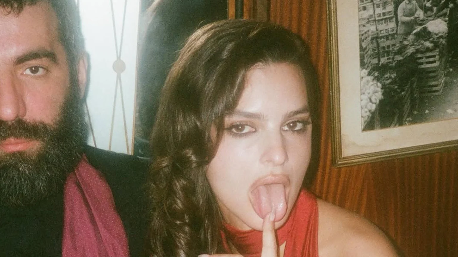 A partially visible man with a dark beard and a red scarf stands next to a woman in a red top, who looks at the camera with her tongue out and a finger touching it.