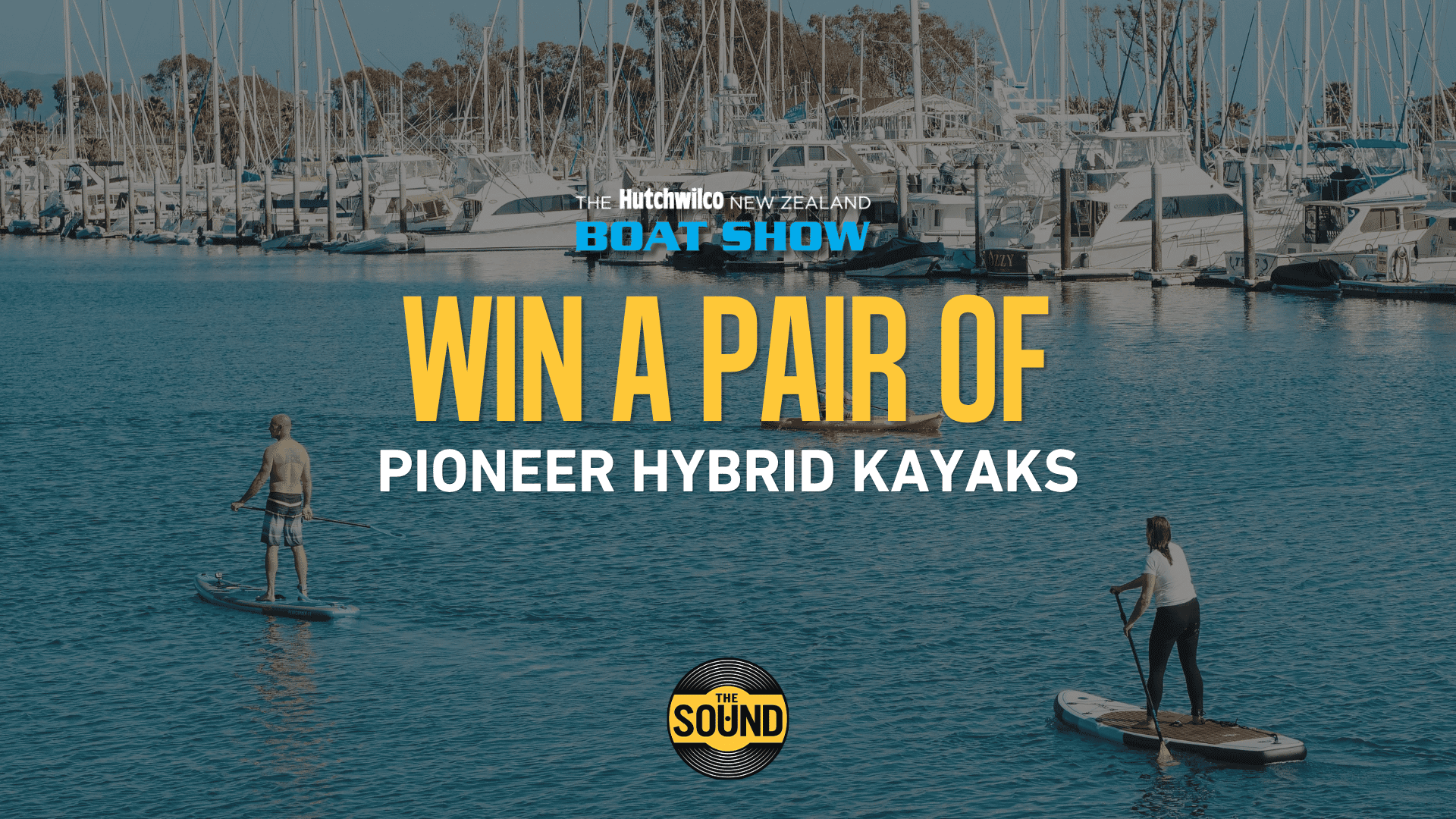 Two people paddleboard on the water in front of a busy marina, with overlay text announcing 'Win A Pair Of Pioneer Hybrid Kayaks' from The Hutchwilco New Zealand Boat Show and The Sound.