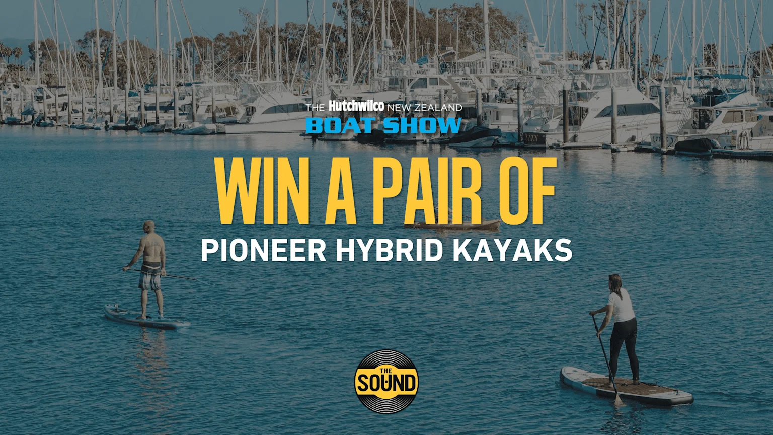 Two people paddleboard on the water in front of a busy marina, with overlay text announcing 'Win A Pair Of Pioneer Hybrid Kayaks' from The Hutchwilco New Zealand Boat Show and The Sound.