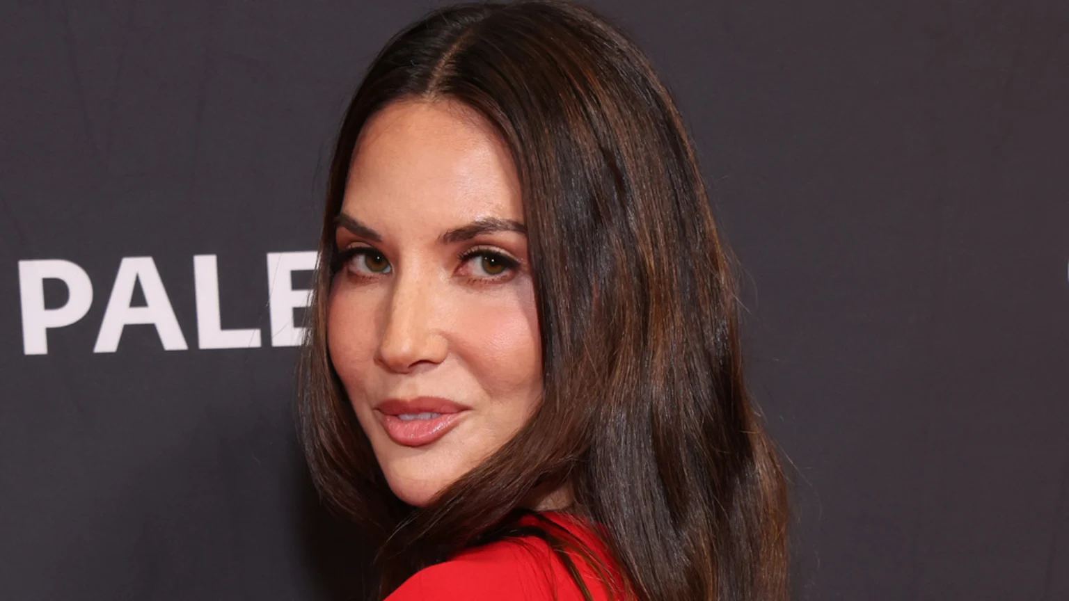 Close-up of Olivia Munn with long dark brown hair and light brown eyes, wearing a red top, looking at the viewer, with the white word 'PALE' partially visible on a dark gray background.