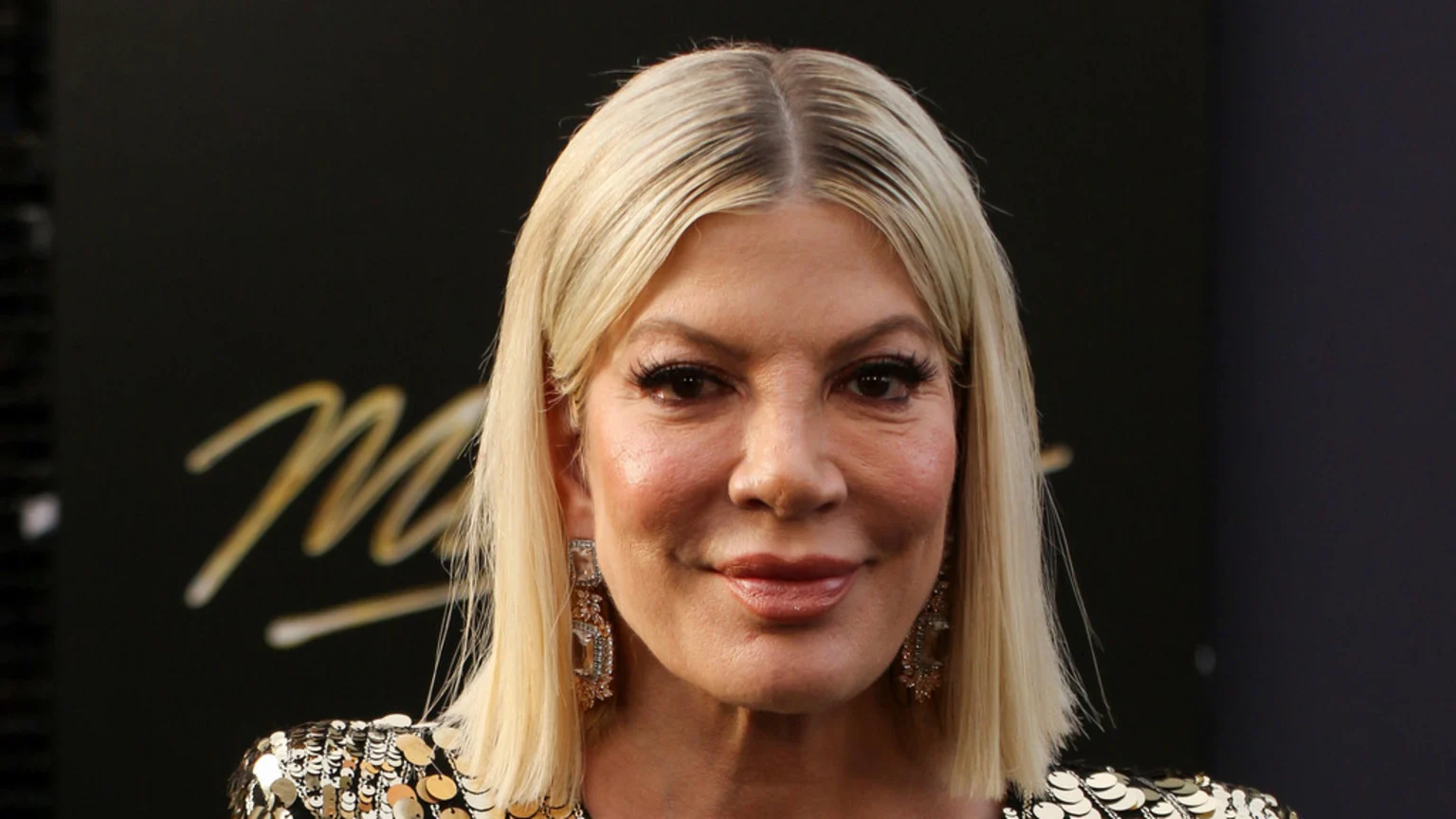 A blonde woman with heavy eye makeup, prominent earrings, and a sequined top smiles slightly, against a dark, blurry background with golden script.