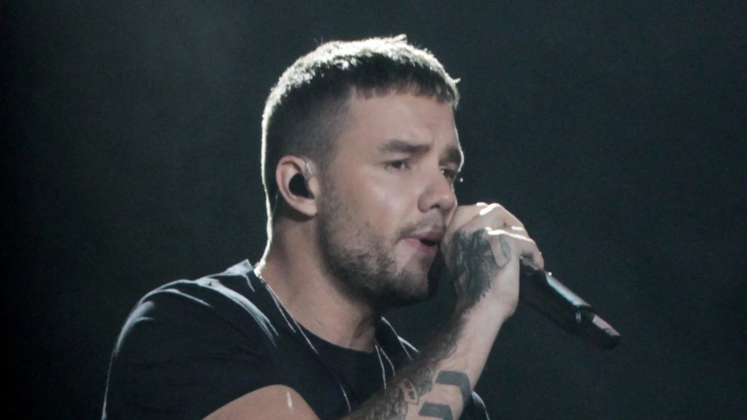 Liam Payne, with short hair and a beard, wears a black t-shirt, holds a microphone with his tattooed right hand, and has an in-ear monitor in his left ear.