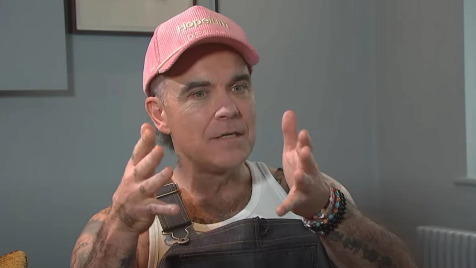 Robbie Williams wearing a pink cap in an interview, gesturing while speaking