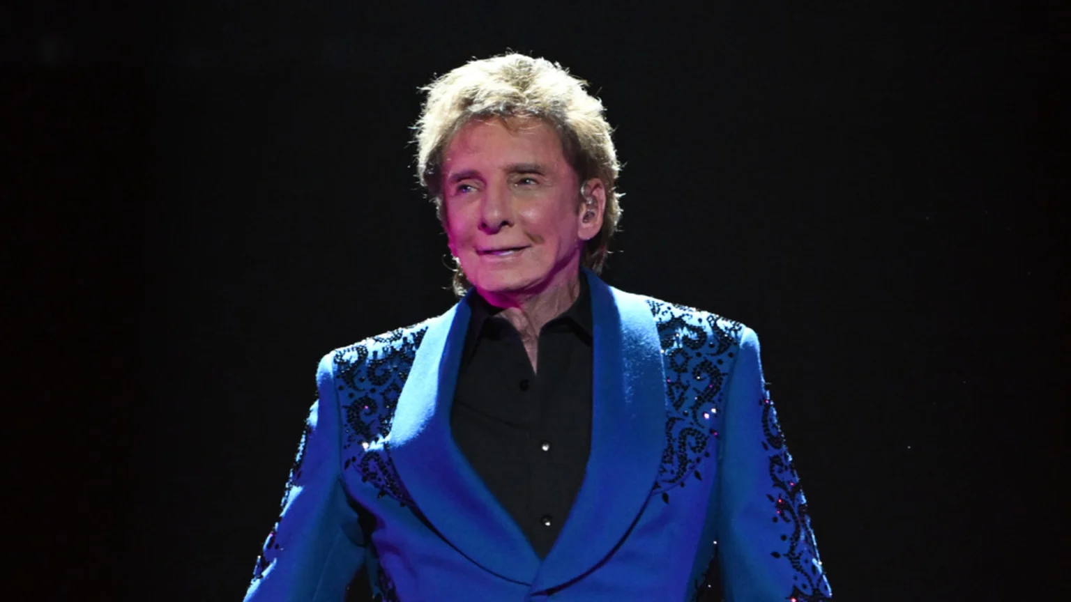 A smiling Barry Manilow with light brown hair wears a bright blue, elaborately decorated jacket over a black shirt, against a dark background.