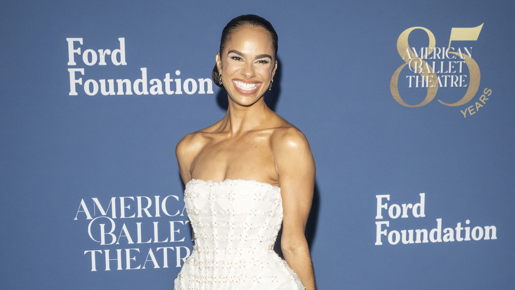 Misty Copeland, in a white strapless dress, smiles broadly against a blue wall with "Ford Foundation" and "American Ballet Theatre 85 YEARS" logos.