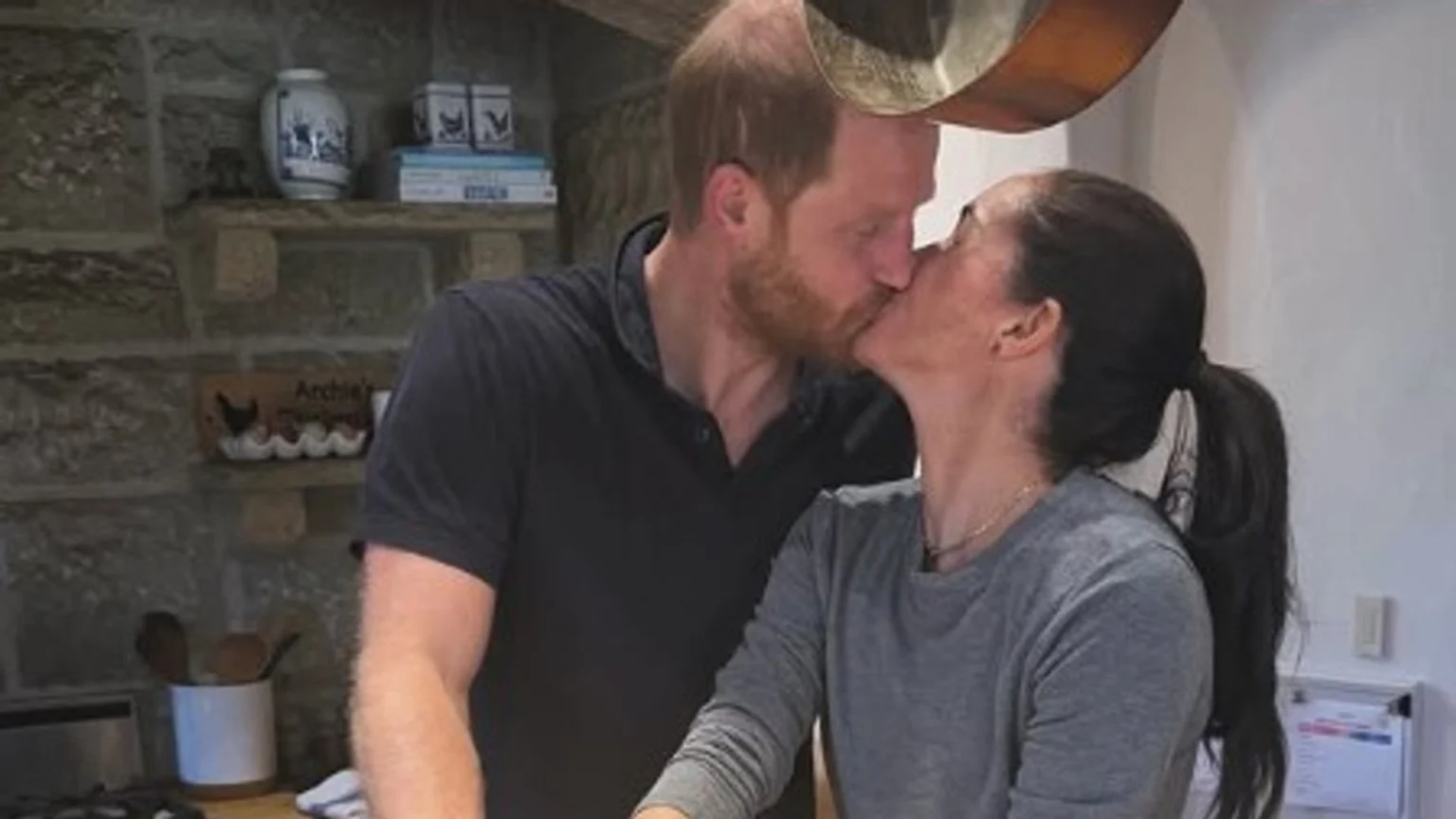 Prince Harry and Meghan Markle kiss in a kitchen with a stone wall background, a shelf holding decor, and a visible sign that reads "Archie's Farm" above an egg carton.