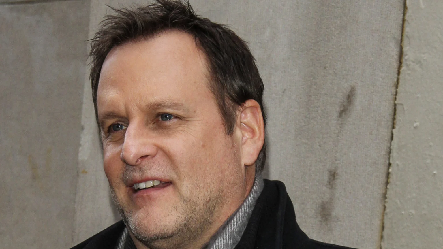 Close-up of actor Dave Coulier, a smiling middle-aged man with short brown hair, blue eyes, and light stubble, wearing a dark coat and grey striped scarf against a light grey wall.