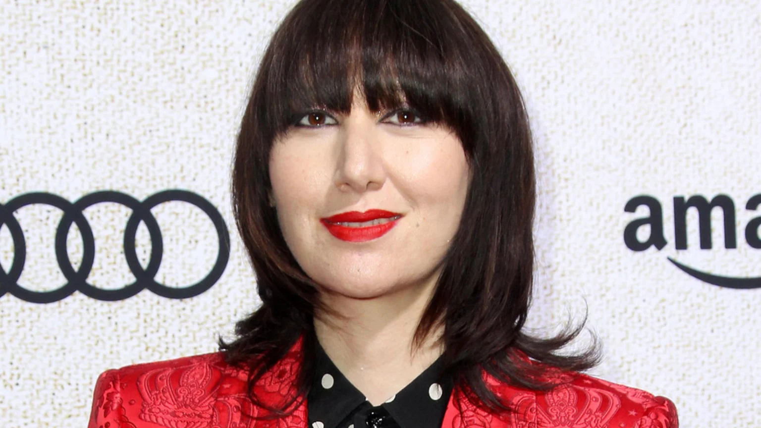 A woman with dark hair and bangs, red lipstick, and a red patterned jacket smiles slightly against a light background with blurred Audi and Amazon logos.