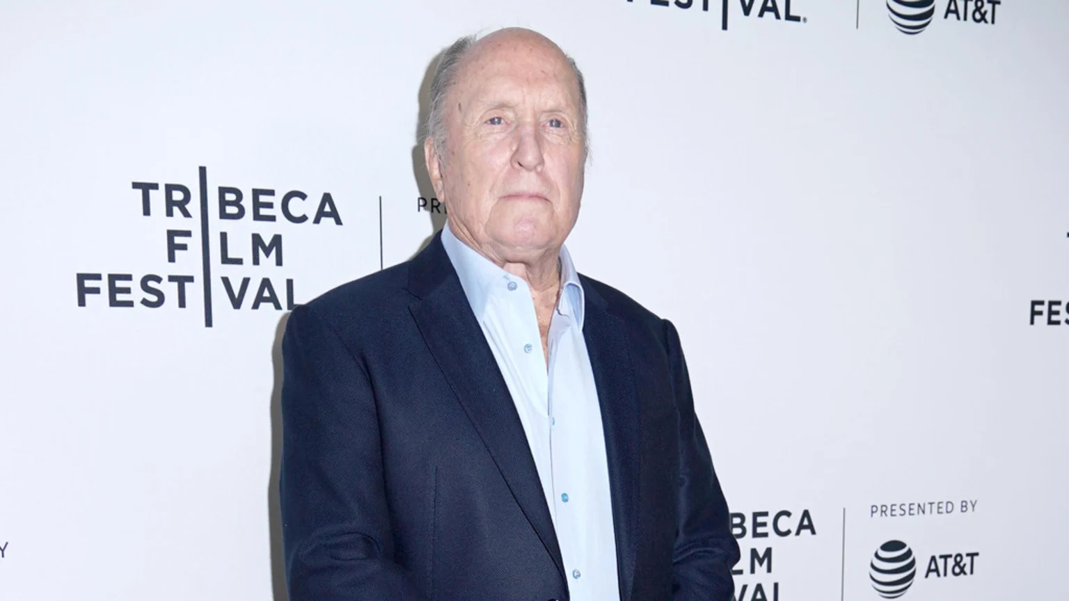 A headshot portrait of Robert Duvall, an older man in a dark blue suit jacket and light blue shirt, standing indoors in front of a white event backdrop with the 'TRIBECA FILM FESTIVAL' and 'AT&T' logos under bright, even lighting.