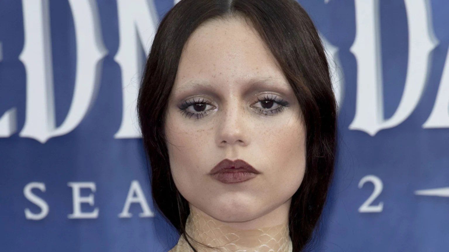 Close-up of Jenna Ortega with dark hair, bleached eyebrows, heavy dark eye makeup, and dark lipstick, posing in front of a blue banner with white text that partially reads "WEDNESDAY SEA 2."