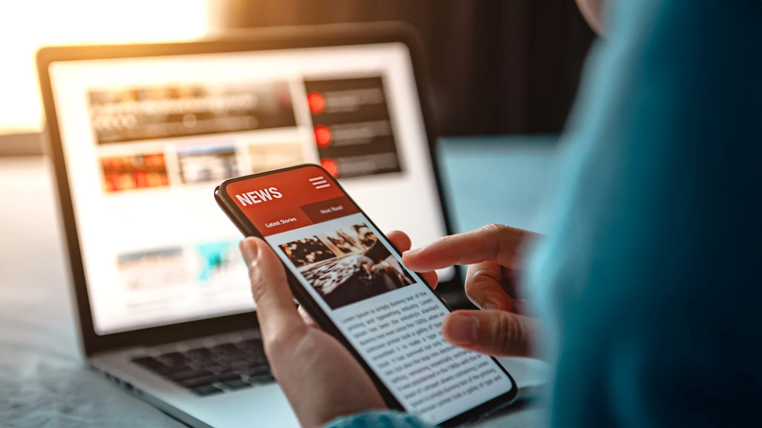 Picture of a person using their phone, with 'news' on it. Blurred picture of open laptop in the background.