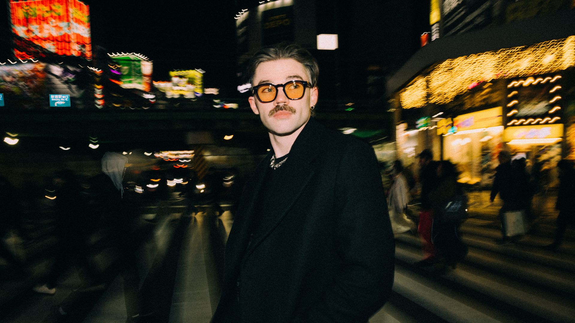 A man with a mustache and orange-tinted sunglasses, wearing a dark coat, stands on a vibrant, blurry city street at night, illuminated by abstract colorful lights and blurred figures.