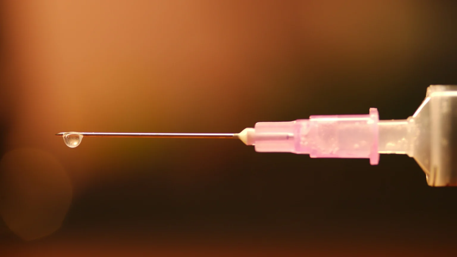 A close-up of a medical syringe needle with a small clear drop of liquid at its tip, set against a blurred warm and dark background.