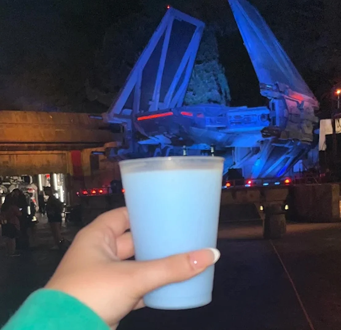 Plastic cup of blue milk held up in front of Disneyland California's lit up Millenium Falcon at night.