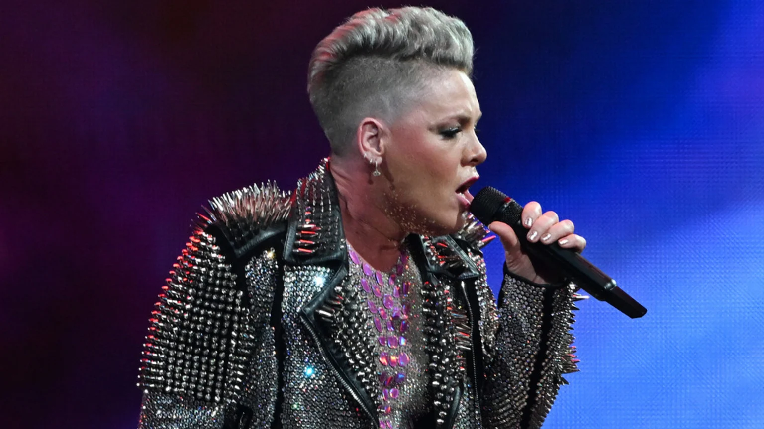 A close-up profile view of Pink singing into a microphone on an indoor concert stage. She wears a black leather jacket with silver spikes and studs, an iridescent purple-pink scale-like top, and a silver dangle earring. The background features dynamic blue and purple stage lighting.