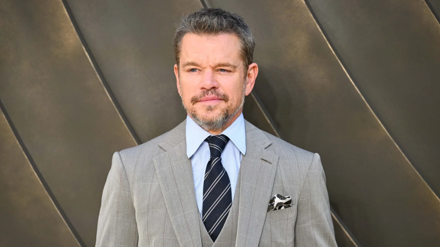 Matt Damon, with grey hair and beard, wears a light grey pinstripe suit, a light blue shirt, and a striped dark tie, looking slightly left against a dark, diagonally patterned wall.