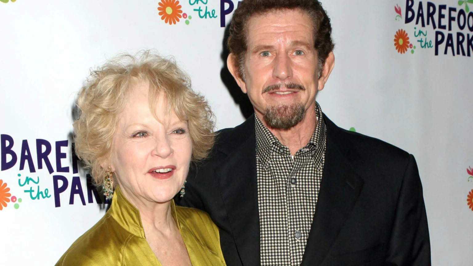 A smiling older woman with curly blonde hair in a gold top stands next to a smiling man with a goatee in a dark suit jacket, posing in front of "Barefoot in the Park" event signage.