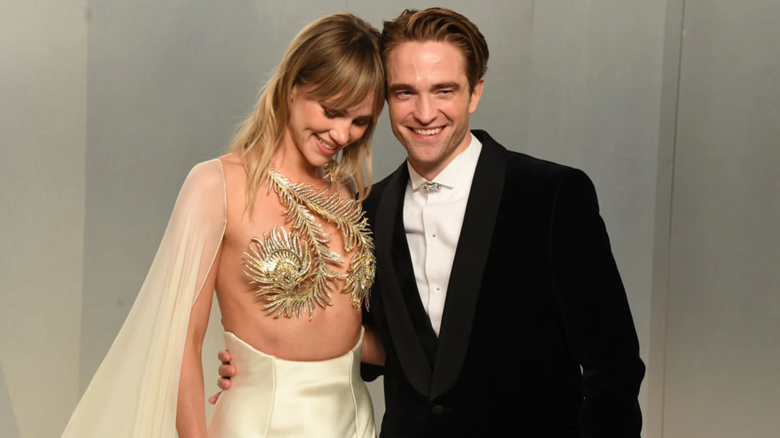 Suki Waterhouse, in a cream dress with gold embellishments and a sheer cape, and Robert Pattinson, in a black tuxedo, stand smiling warmly together.