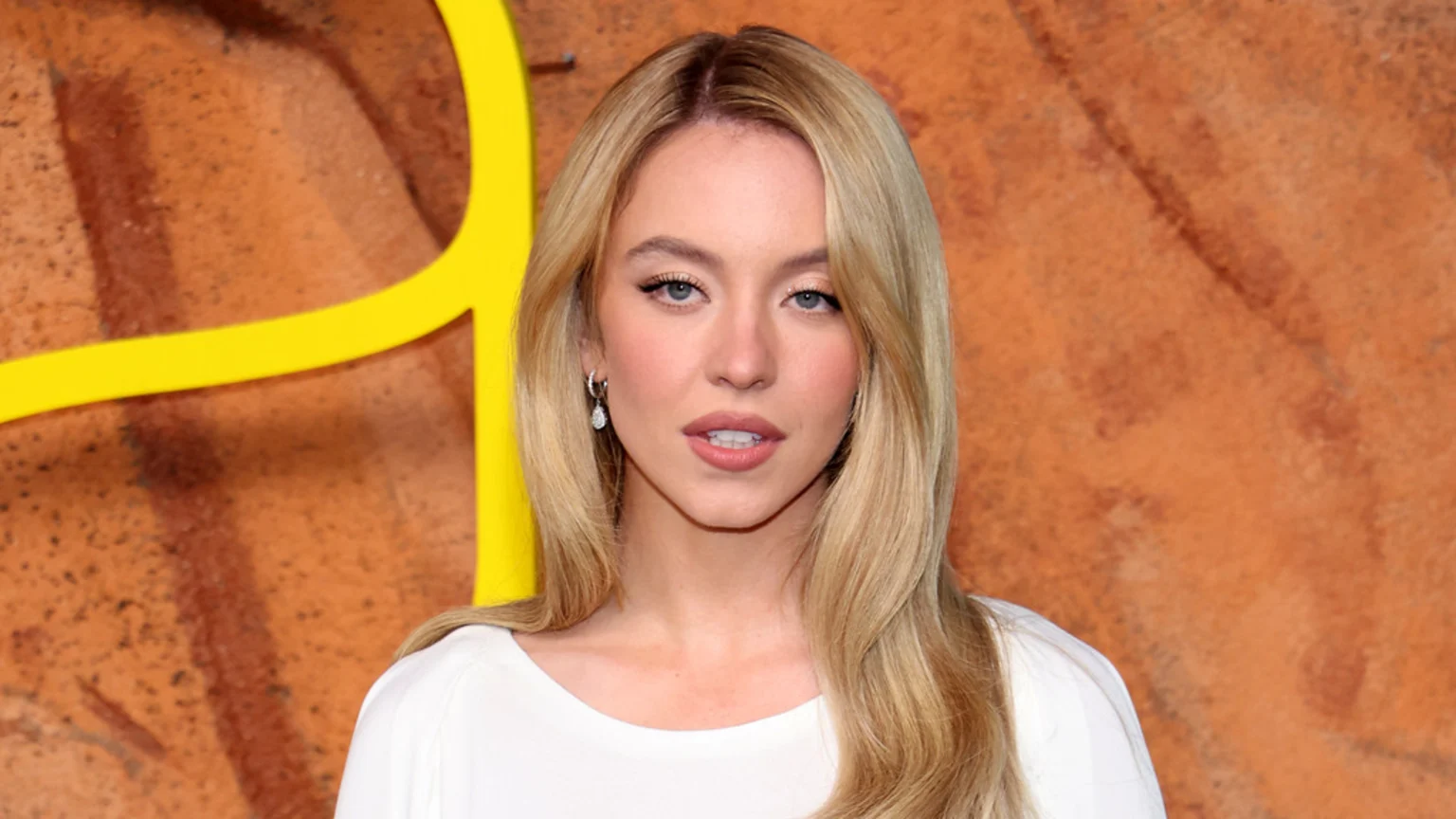 Sydney Sweeney, a blonde woman, looks directly at the camera wearing a white top, with an orange and yellow background.