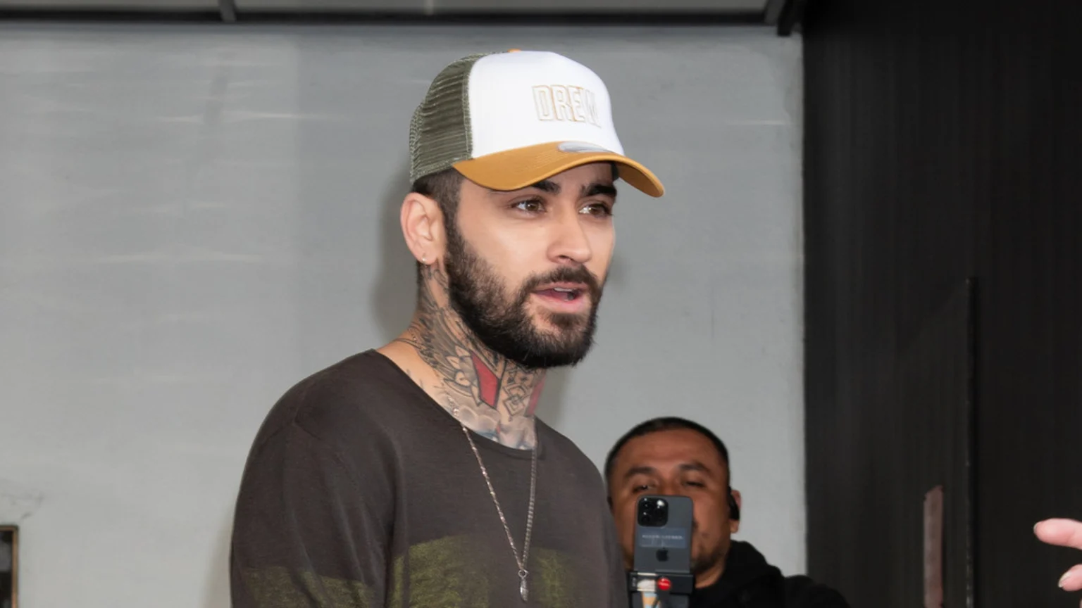 A man with a beard and neck tattoos wears a white and olive green "DREW" trucker hat and a dark shirt, speaking, with another person recording in the background.