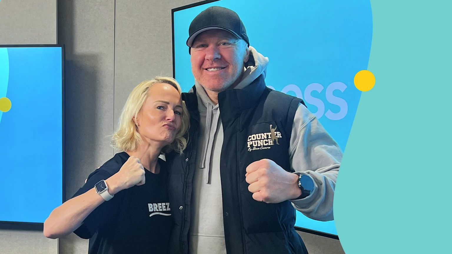 A man in a black cap, grey hoodie, and black vest with "COUNTER PUNCH By Shane Cameron" logo smiles and makes a fist while standing next to a blonde woman making a playful pouting face and also making a fist, in front of a blue screen.