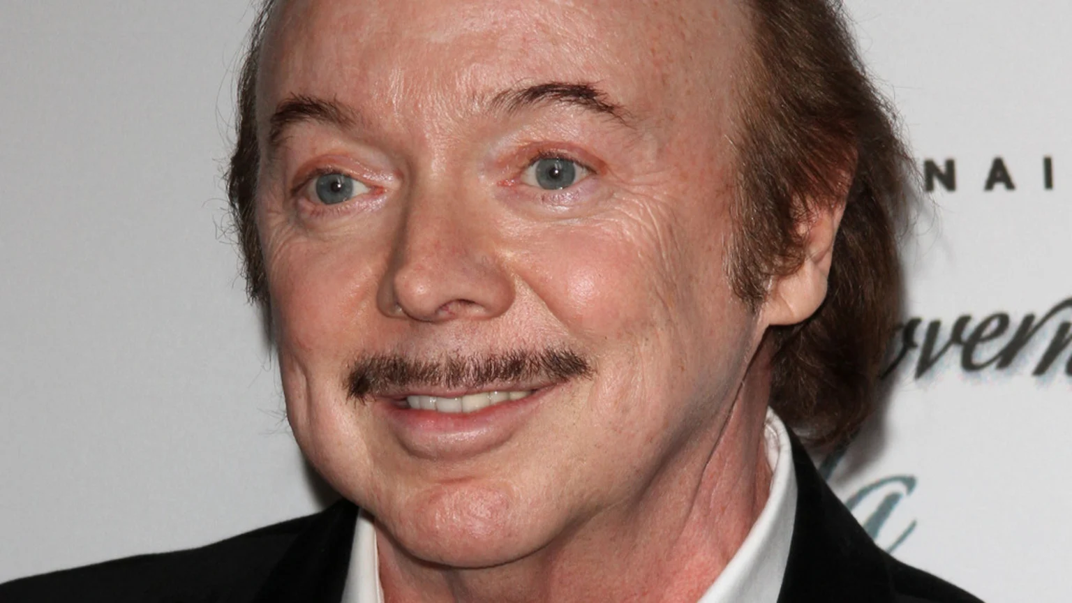 A close-up, head-and-shoulders portrait of an older man with brown hair and a prominent mustache smiling slightly. He wears a dark suit jacket and white shirt, posed under bright even lighting against a light background with partial text signage visible.
