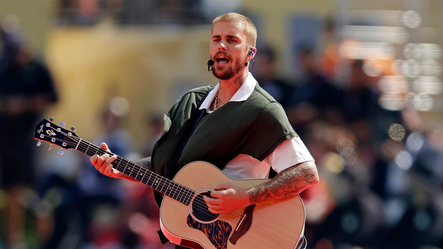 Justin Bieber, with a beard and arm tattoos, sings into a microphone while playing an acoustic guitar, wearing a dark green top over a white shirt.