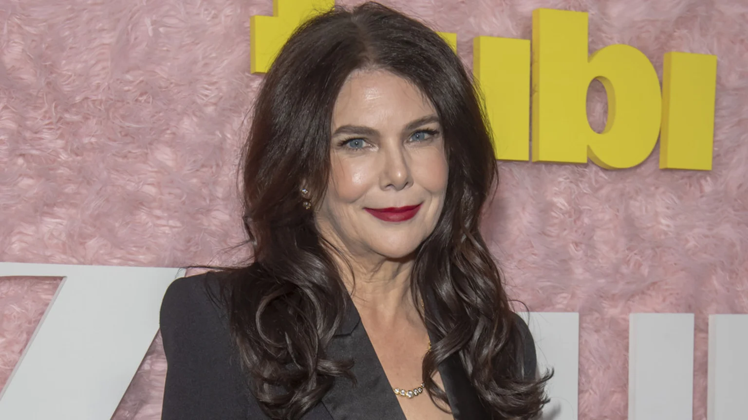 Actress Lauren Graham with dark wavy hair and blue eyes, wearing a black blazer, red lipstick, and diamond jewelry, smiles against a pink fluffy background with yellow "Tubi" branding.