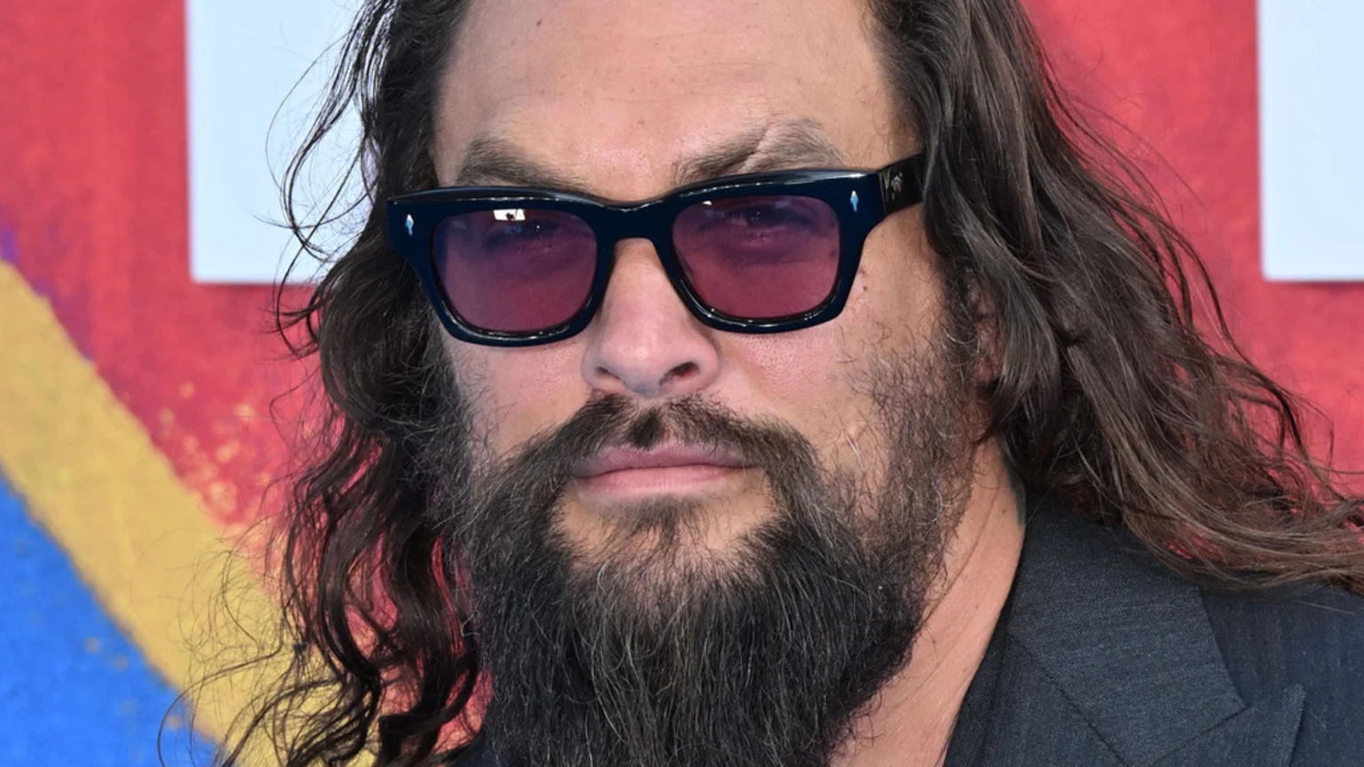 Close-up of actor Jason Momoa, facing forward, with long dark hair, a full beard, and a prominent scar above his left eyebrow, wearing dark sunglasses with purple lenses against a vibrant, abstract background.