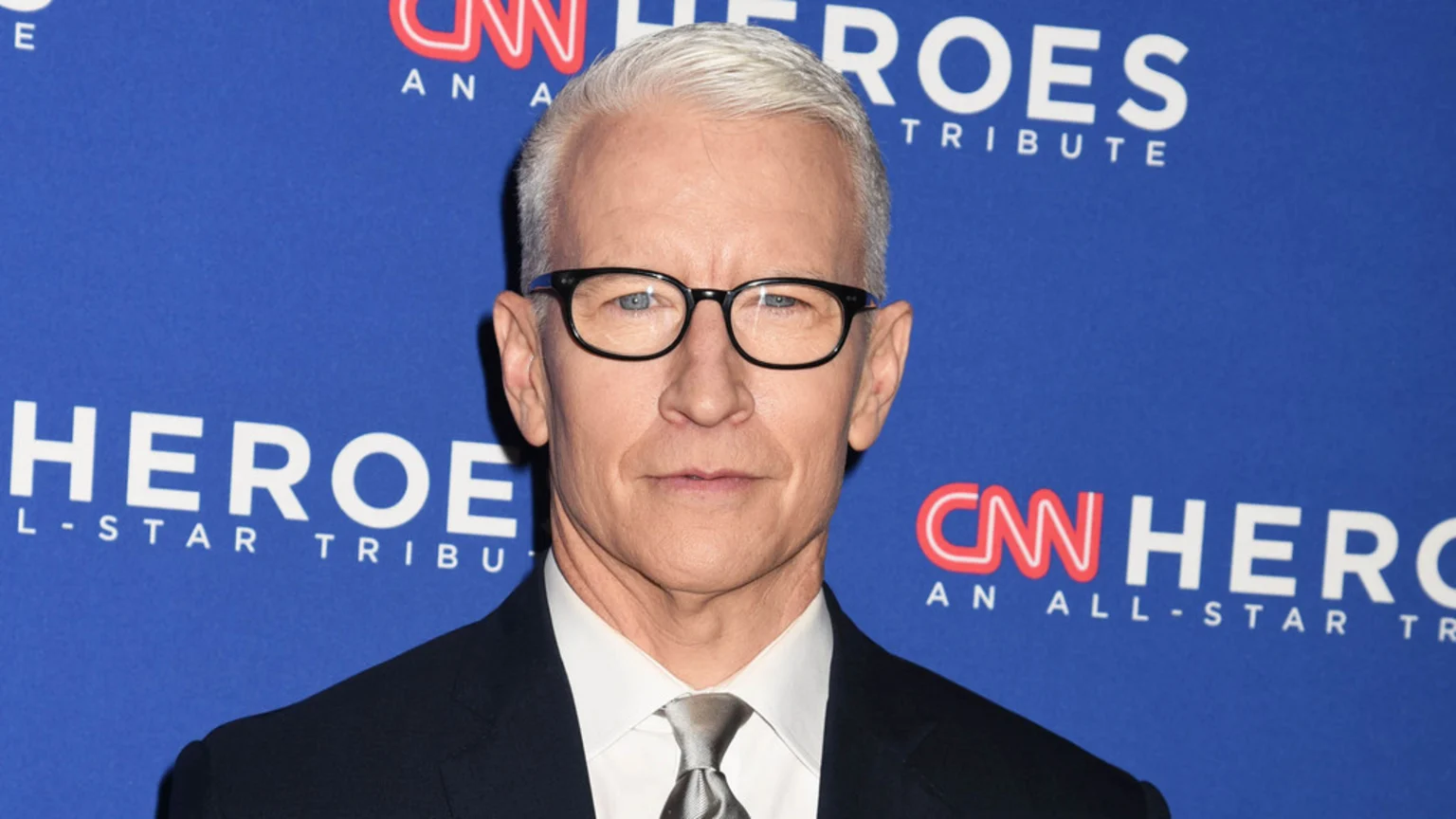 A close-up portrait of Anderson Cooper wearing glasses, a dark suit, white shirt, and gray tie, standing against a blue backdrop with repeated 'CNN HEROES AN ALL-STAR TRIBUTE' text under bright studio lighting.