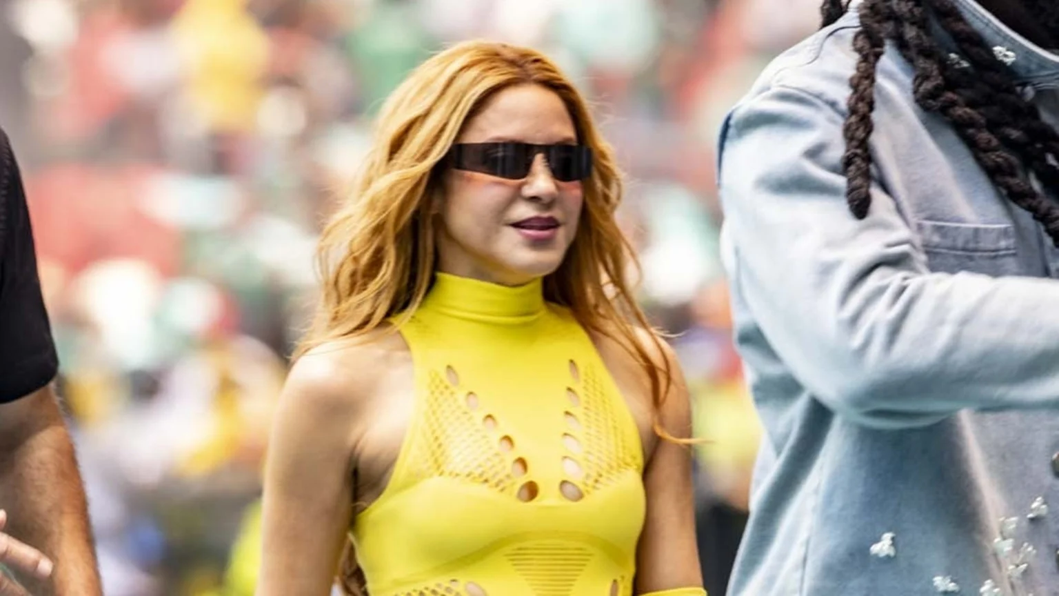 Shakira, with long wavy blonde-red hair, wears a bright yellow sleeveless top with cutouts and dark sunglasses, standing beside a man with dreadlocks and another partially seen person.