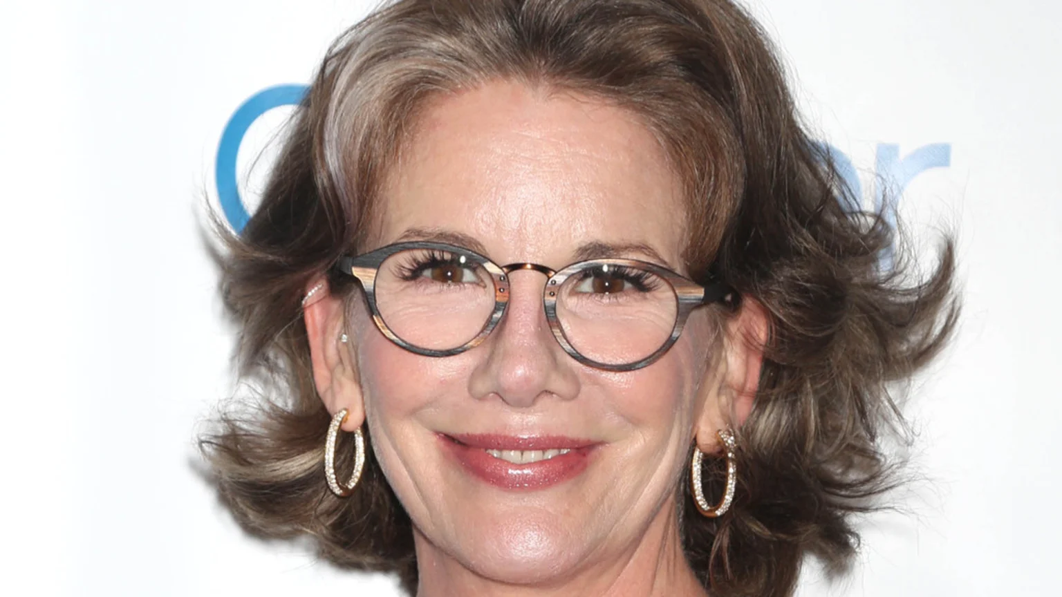 Close-up of Melissa Gilbert smiling, wearing distinctive wood-patterned glasses, gold hoop earrings, and glossy pink lipstick, with her wavy brown and gray hair visible.