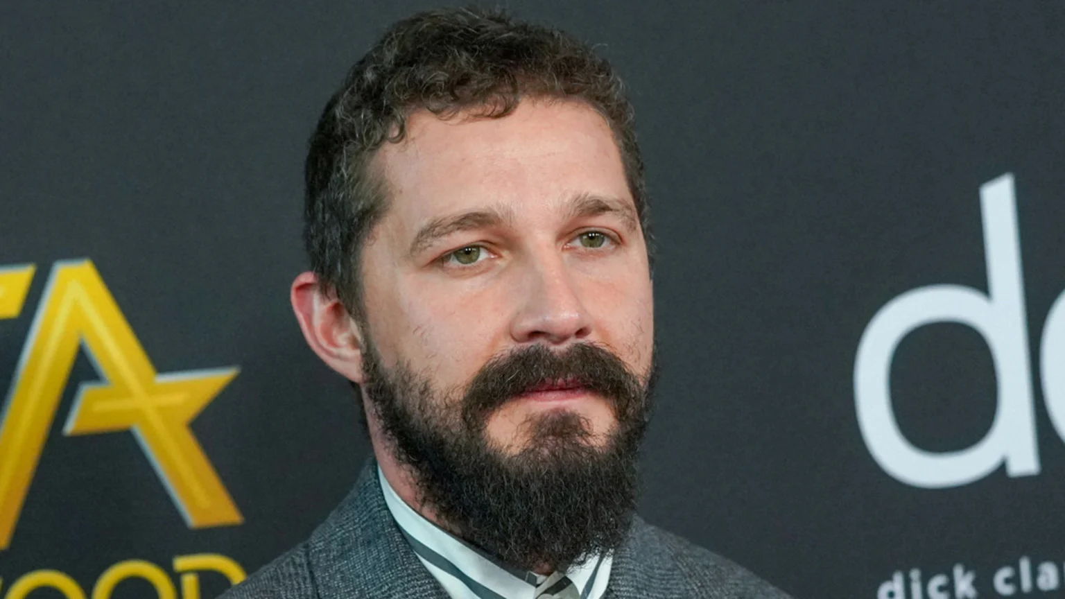 Shia LaBeouf, with curly hair, a beard, and green eyes, looks right while wearing a suit against a dark background with blurred logos.