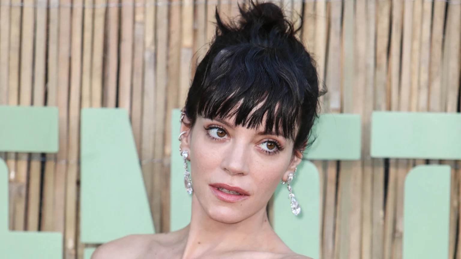 A woman with dark hair in a messy bun and bangs wears multiple earrings, including long dangling ones, looking upwards with a slight smirk against a blurred background of wooden slats and light green shapes.