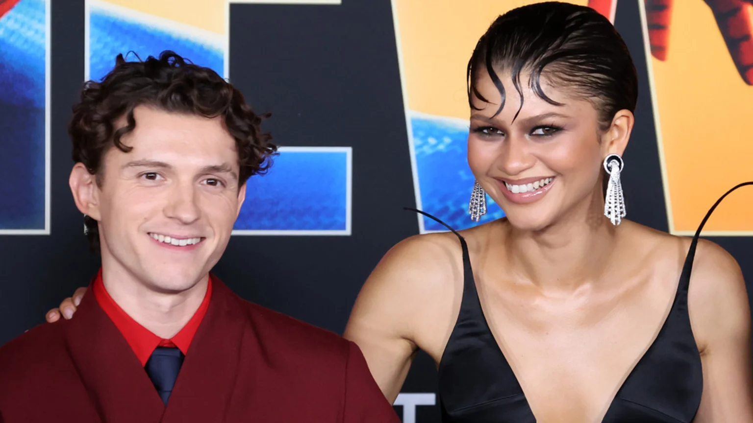 Tom Holland and Zendaya pose together, smiling widely at the camera, with Tom in a red suit and Zendaya in a black dress, her arm around him.