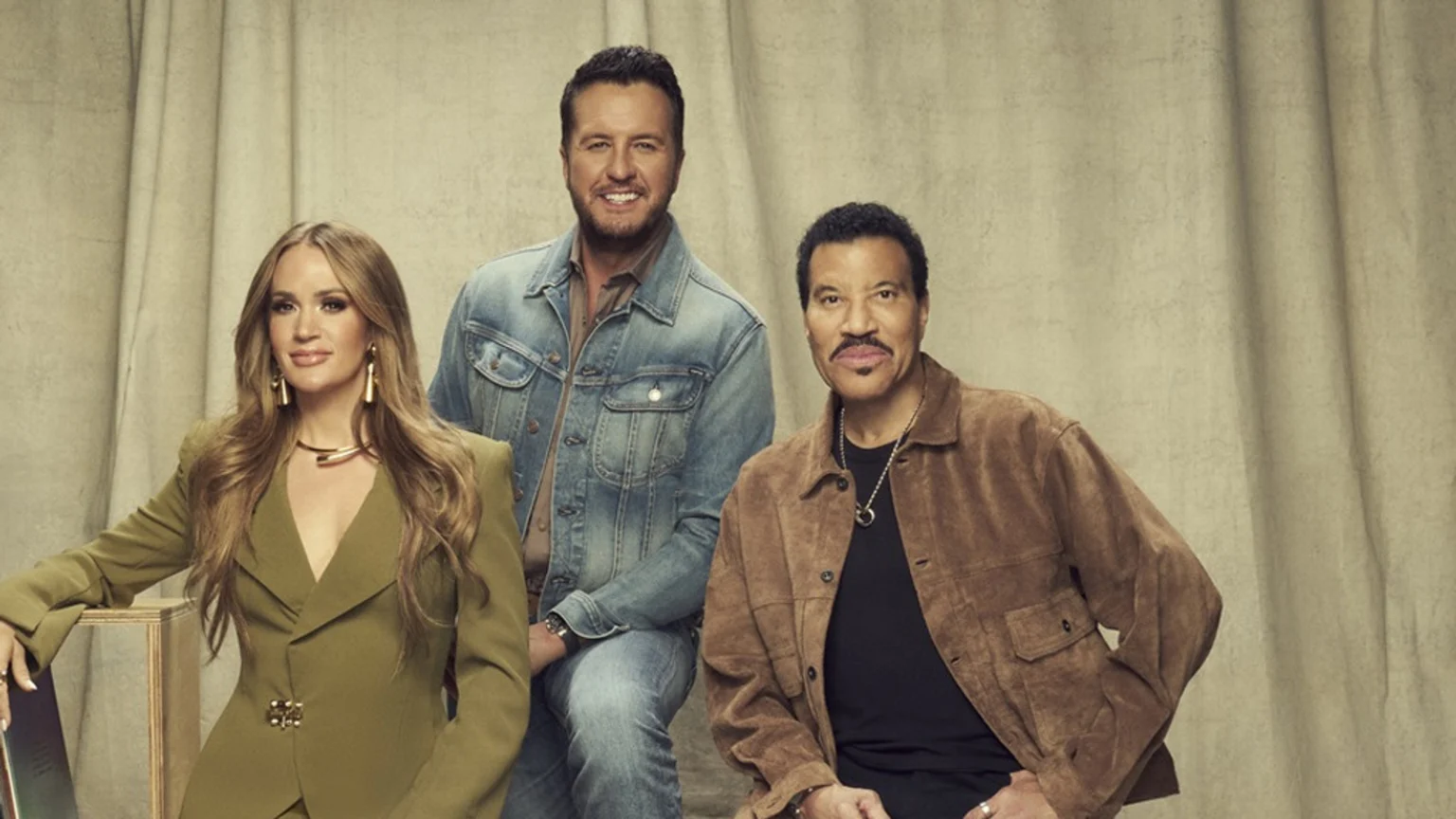 Carrie Underwood, Luke Bryan, and Lionel Richie are seated, smiling at the camera. Underwood wears an olive suit, Bryan a denim jacket, and Richie a brown suede jacket, against a beige studio backdrop.